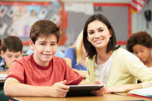 Pupils In Class Using Digital Tablet With Teacher Smiling At Camera